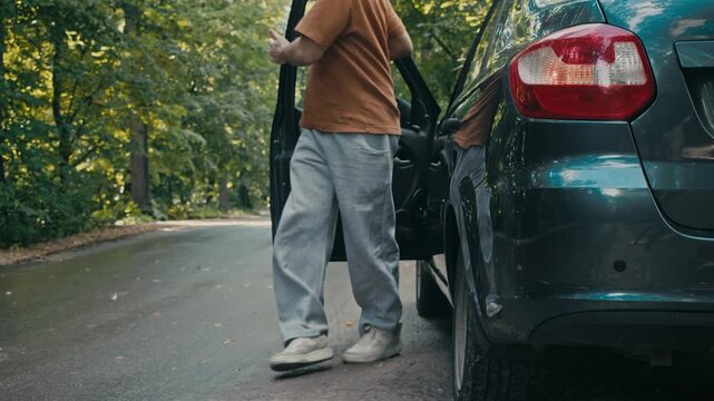 Driver crouches near rear wheel checking damaged tire on roadside. Car stands in forest with hazard lights flashing after breakdown.