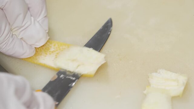 Close up of a professional pastry chef wearing white latex gloves carefully trimming white pith from a fresh lemon peel with a sharp knife on a plastic board. Prepared for gourmet dessert decoration, 