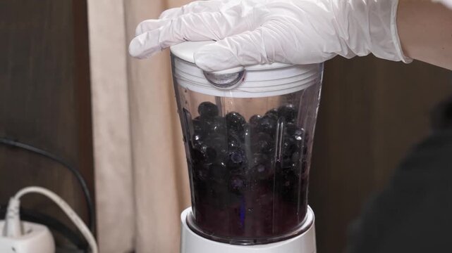 close-up shot of a person wearing a white disposable glove placing the lid onto a clear blender jar packed with fresh, dark blueberries, preparing to blend a healthy fruit dessert or smoothie.