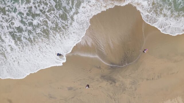 Aerial 4K video looking straight down at golden sand as gentle ocean waves roll onto the beach, creating white foam before slowly receding in bright midday sunlight.