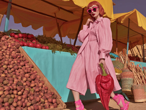 Woman in a pink coat and sunglasses walking beside a large display of potatoes. Perfect for fashion editorials, accessory campaigns, travel branding, and retail advertising.