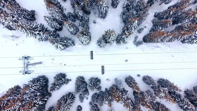 Top down aerial view of ski chairlift. Two ski lift cabins travel over white snow between rows of evergreen pine trees.