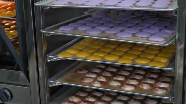 A professional baker wearing white gloves slides a baking sheet filled with bright orange macaron halves into a commercial rack oven. Other trays containing purple and pink macaron shells are visible
