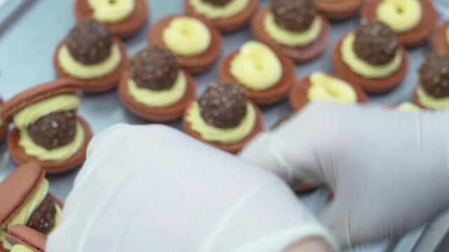 close-up shot of a pastry chef wearing white gloves carefully assembling gourmet chocolate macarons on a metal tray. Each macaron shell is filled with a swirl of yellow buttercream frosting and topped