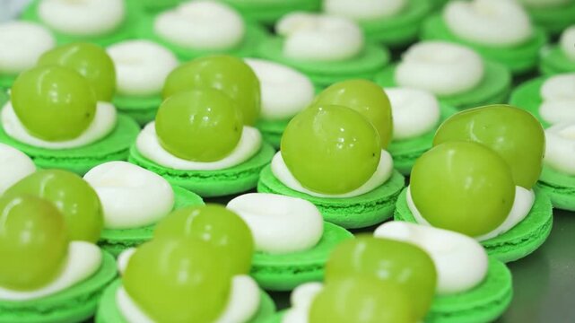 close-up high-angle shot showing rows of gourmet green French macarons being assembled. Each bottom shell is topped with a smooth swirl of white whipped cream or ganache and garnished with fresh, juic