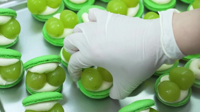 close-up shot of a pastry chef wearing white gloves, carefully assembling vibrant green macaron cookies filled with fresh green grapes and smooth white pastry cream on a commercial kitchen tray