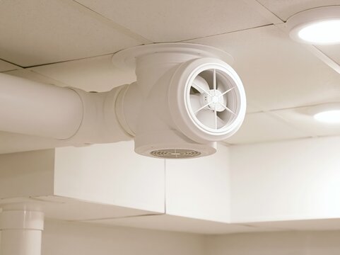 A white ventilation fan is mounted on a pipe in a room with white ceiling.