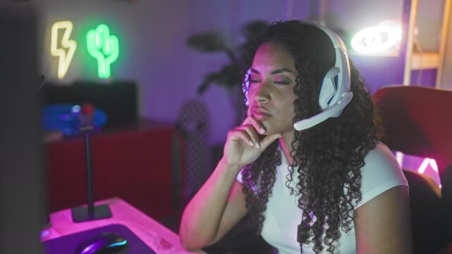 Woman with curly hair wearing a headset gestures with fingers while seated at a computer in a gaming studio with neon signs, rgb lighting and a red gaming chair; thoughtful focus.