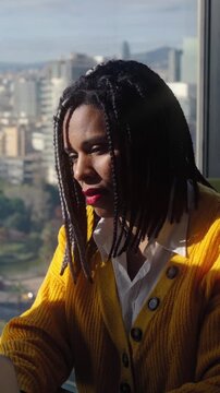 Vertical. Focused Black businesswoman working on a laptop in a modern high-rise office. Sunlit corporate scene with panoramic city views conveys concentration, leadership and professional
