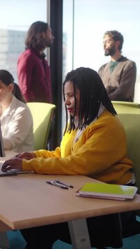 Vertical. Focused african millennial woman in yellow sweater using laptop in a bright office with colleagues, teamwork atmosphere, modern business workspace, city view