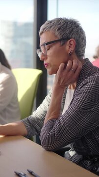 Vertical. Middle aged professional works at an open plan desk in a bright corporate office, concentrating on tasks at a dual monitor setup with paperwork and notes by the window overlooking the city.