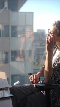 Vertical. Focused mature businesswoman typing on a laptop at a spacious desk beside panoramic office windows. Wide corporate scene conveying productivity, concentration and professional independence.