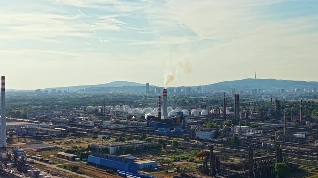 Factories are active in the foreground with smokestacks emitting smoke while a city skyline and mountains are visible in the background during the afternoon sunlight.