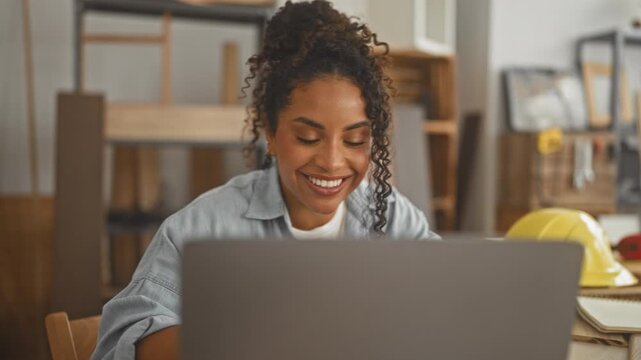 Woman carpenter smiles at laptop with yellow hardhat and wooden boards in a workshop; confidence creativity.