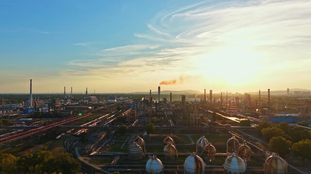 A skyline view of an industrial area during sunset shows tall chimneys and round storage tanks. Smoke rises into the air, creating a busy atmosphere.
