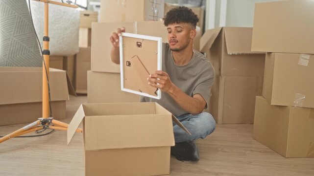 Man holding picture frame with both hands while sitting among moving boxes in building; moving day curiosity.