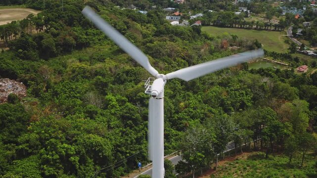 Close up wind turbine spinning blades over Phuket's green landscape, generating clean renewable energy beside roads and homes, symbolizing sustainable infrastructure and climate action. Drone shot