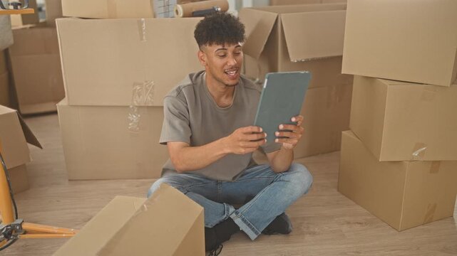 Man holding tablet, hand to neck while swiping and gesturing among stacked cardboard moving boxes in building; focused planning.