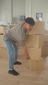 Man grimacing and lifting stacked cardboard boxes, leans awkwardly and holds lower back for backache in building; pain struggle.