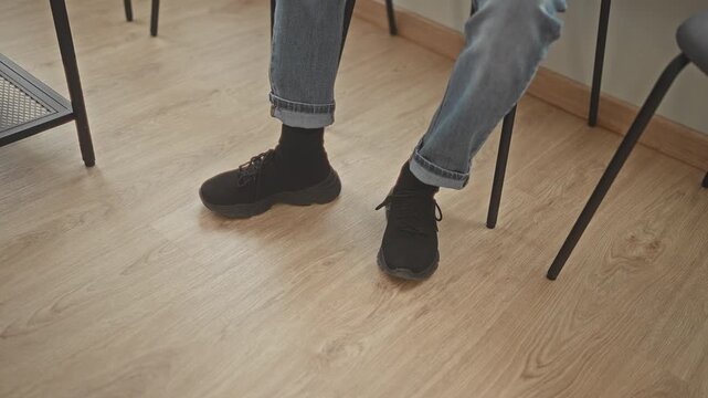 Man sitting on chair with legs crossed wearing black sneakers on wooden floor, waiting in studio; quiet anticipation.
