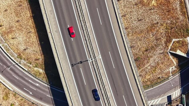 A car moves along the highway under an overpass. The road is surrounded by dry land and infrastructure. It is bright and clear outside, likely in the afternoon.