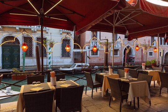 Outdoor restaurant terrace beside a narrow Venetian canal with tables, boats and traditional architecture in Venice, Italy.
