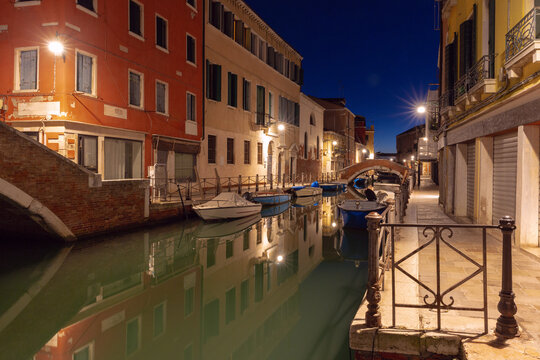 Quiet canal with moored boats, brick bridges and colorful buildings reflected in calm water before sunrise in Venice, Italy.