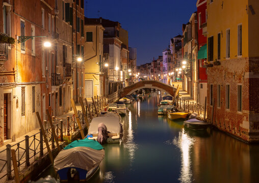 Quiet canal with moored boats, brick bridges and colorful buildings reflected in calm water before sunrise in Venice, Italy.
