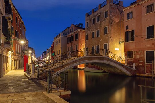 Quiet canal with moored boats, brick bridges and colorful buildings reflected in calm water before sunrise in Venice, Italy.