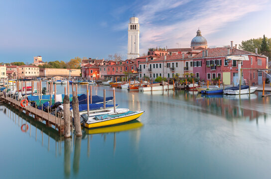 Boats moored along wooden piers beside traditional buildings and a bell tower at dawn in the Castello district of Venice, Italy.