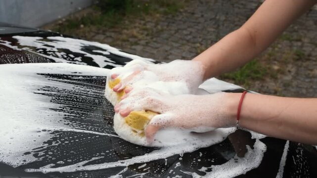 Person washing a black car with both hands using a soapy sponge, covering the surface with foam during careful vehicle cleaning and automotive detailing