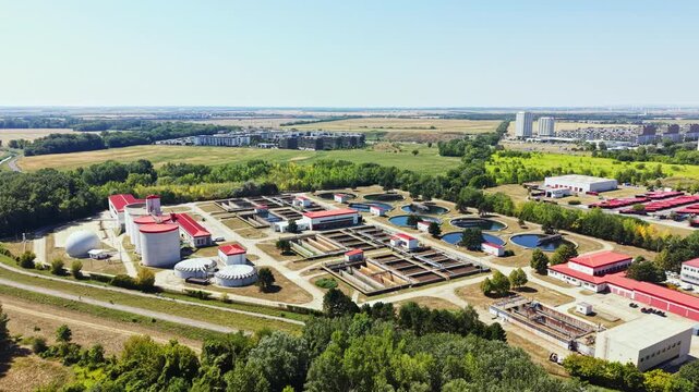 Workers are processing water at a wastewater treatment plant near a city during daytime. The field surrounds the plant, with buildings seen in the distance.