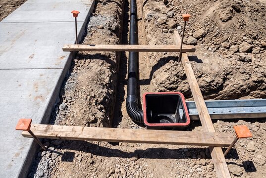 Black corrugated drainage pipe and catch basin installed in a dirt trench next to a concrete sidewalk