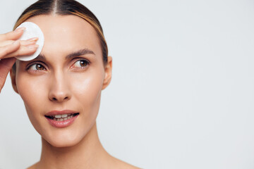 Young woman with brunette hair gently cleanses her face using a cotton pad against a plain light background, showcasing a skincare routine with a calm and focused expression. © SHOTPRIME STUDIO