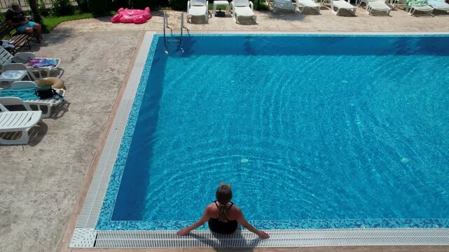 woman sitting on the edge of the pool in hotel from above view, aerial top view from drone, summer relaxation in hotel

