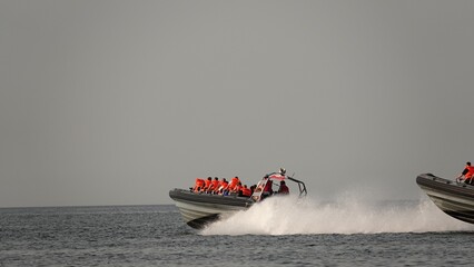 Szybka łódź motorowa typu RIB pełna pasażerów w pomarańczowych kamizelkach ratunkowych przecina morskie fale. Uniesiony dziób jednostki i piana wodna podkreślają dynamikę i dużą prędkość rejsu. © tomo81