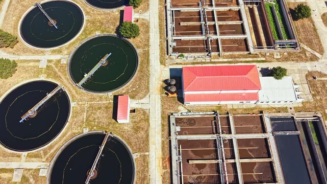 Aerial view of a water treatment plant showing large circular tanks filled with dark water. Nearby are rectangular structures and a building with a red roof. The area is dry and sunny.
