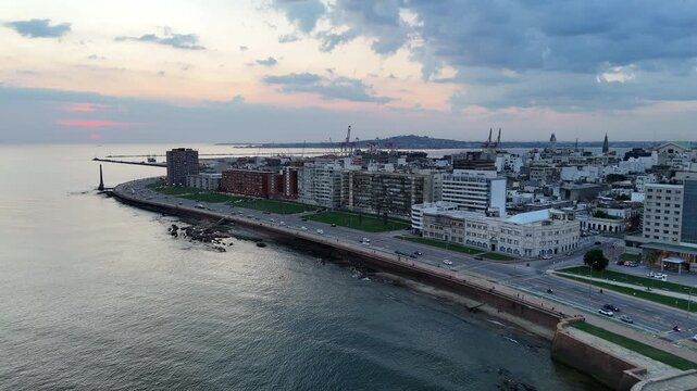 Drone view along Montevideo&rsquo;s waterfront boulevard at dusk, showing coastal traffic, promenade, urban buildings, green spaces and the Port of Montevideo in the distance.