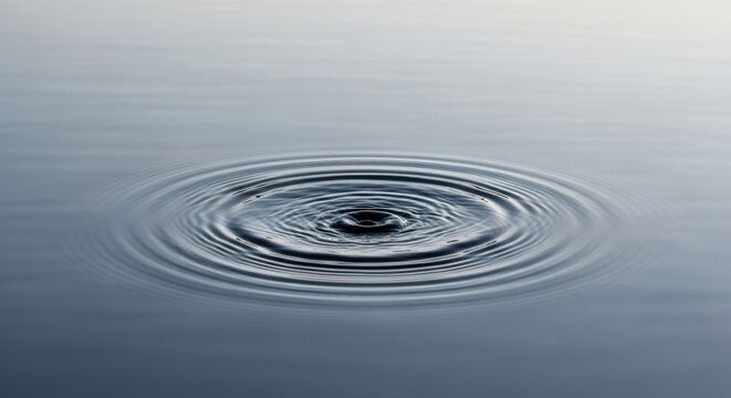 Macro view of concentric water ripples expanding from a central impact point on a smooth and calm dark grey water surface with soft light and circular wave patterns.