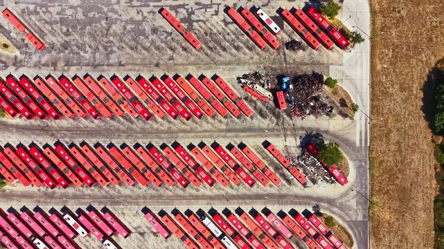 Rows of red buses fill a big parking lot. Some buses are lined up neatly while others are in groups. Nearby, there is a pile of scrap material. The scene appears to be during daytime.