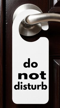 White do not disturb sign hangs on dark wood door near metal handle. Blank notification card signals privacy needed inside hotel room or office space.