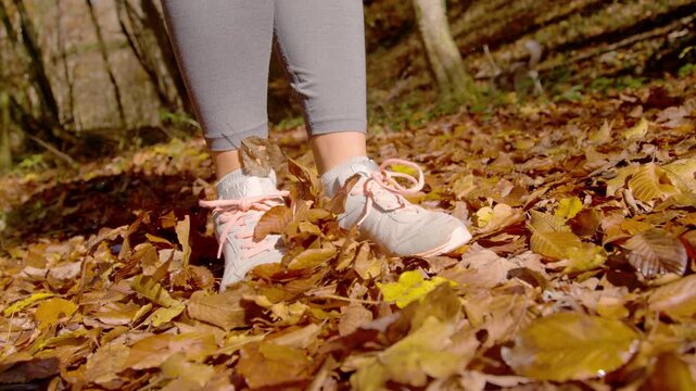 CLOSE UP, LOW ANGLE, SLOW MOTION: Woman stepping through crisp autumn leaves in the sunny forest. Her shoes kick and scatter fallen foliage around. Moment of walking outdoors in colourful fall nature.