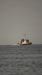 Statek roboczy, przypominający holownik, płynie po spokojnej, lekko pofalowanej tafli morza. Jednostka znajduje się daleko od brzegu, odcinając się na linii horyzontu pod szarym niebem. © tomo81