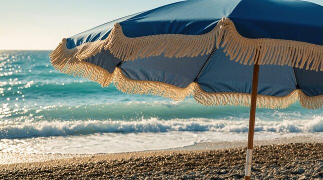 Blue beach umbrella with fringe waves on pebble shore. Ocean water sparkles under sun. Gentle breeze moves fabric. Peaceful seaside vacation scene.