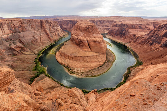 Scenic desert canyon landscape featuring a winding river surrounded by dramatic red rock cliffs, rugged terrain, and layered formations under a soft cloudy sky in the American Southwest.