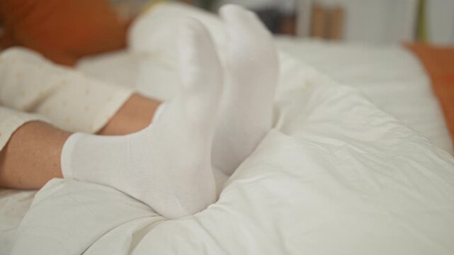 Woman in polka dot pajamas wiggling feet in white ankle socks on a rumpled bed in a bright bedroom; relaxed cozy playfulness.