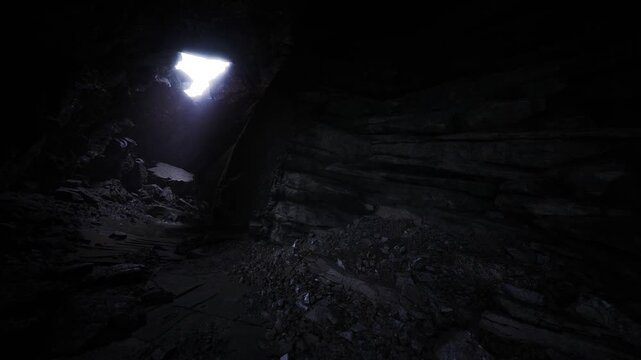Dim chamber with overhead aperture casting diffuse light across rubble and slick stone, eerie stillness, tactile rock detail and low key color