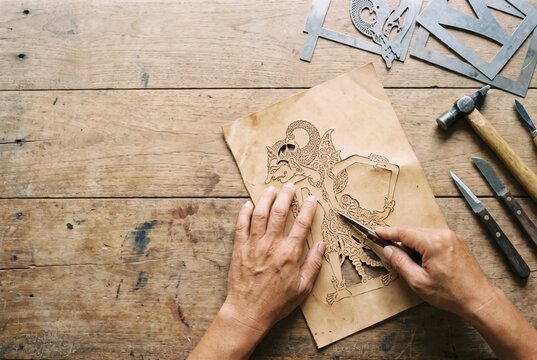 Hands cutting leather shadow puppet outline on wooden craft table