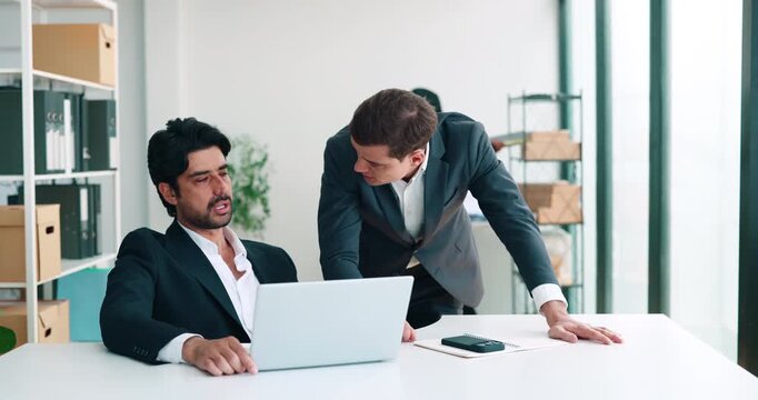 Male boss points at laptop as seated businessman studies screen. Middle eastern colleague reviews project changes following instructions during office workday