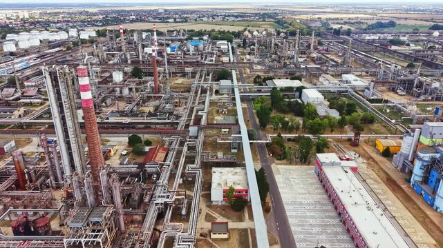 A view of a large industrial area showing multiple factories and pipelines. The sky is clear. The land is flat with some trees and buildings scattered among the structures.